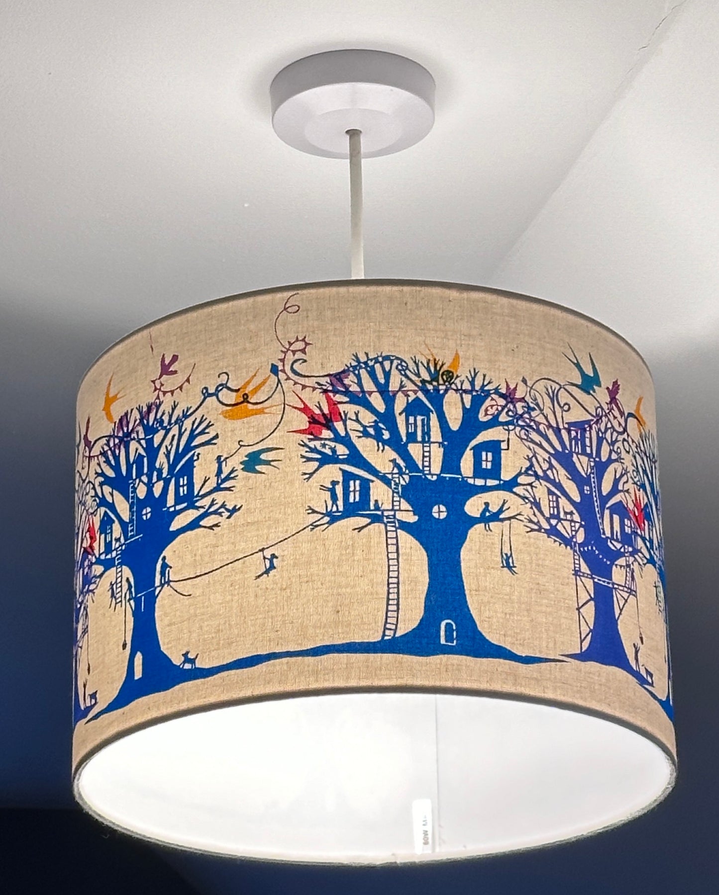 Ceiling lampshade (lit) with blue and turquoise  trees with treehouses on cream background with multi coloured birds and bunting.