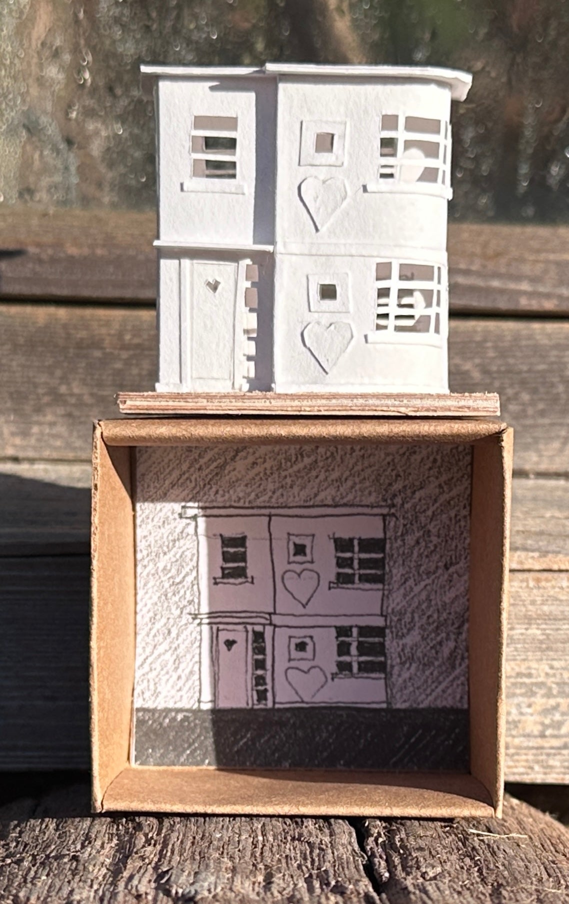 Modern House front elevation of white paper modernist style house with applied hearts on plywood base, standing on presentation box showing pencil drawing on back face.