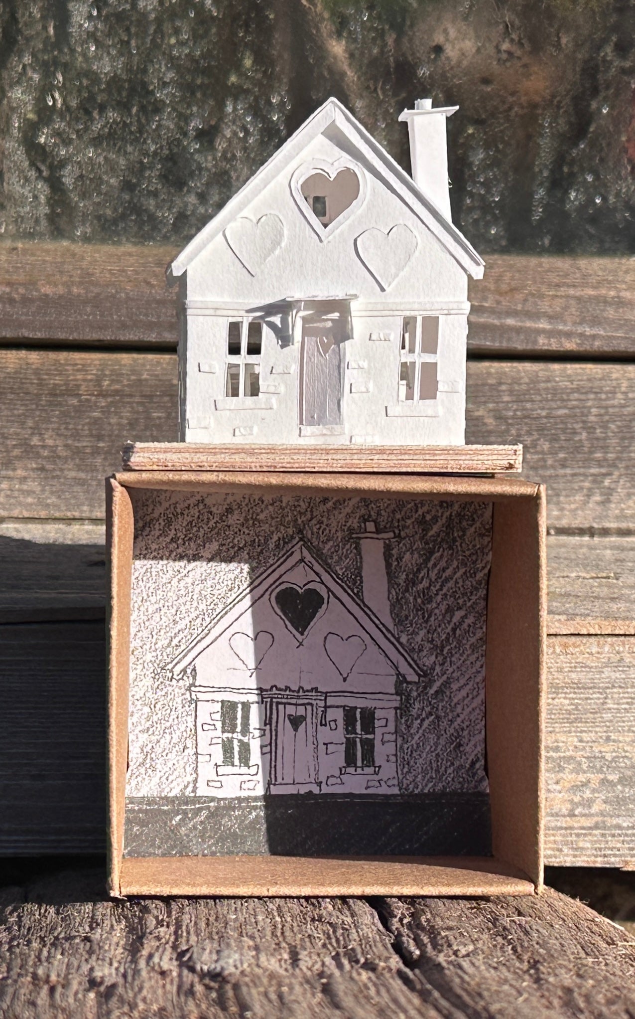 White paper gable fronted house with a heart shaped window and two applied hearts and a  tall chimney, standing on presentation box.