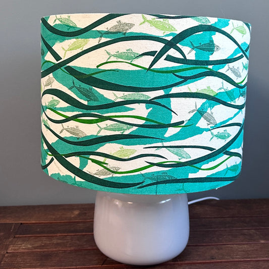 A table mounted lampshade with a  variety of different shades of green fish swim amongst green weeds over a paler green monoprinted background on a natural coloured fabric.