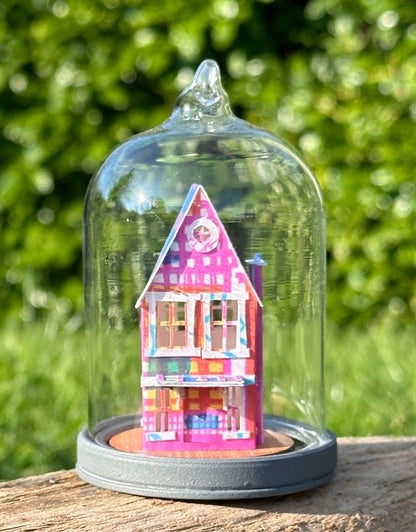 Front elevation of gable roofed house made from multicoloured paper , on a grey wooden base under a glass dome with pigtail top