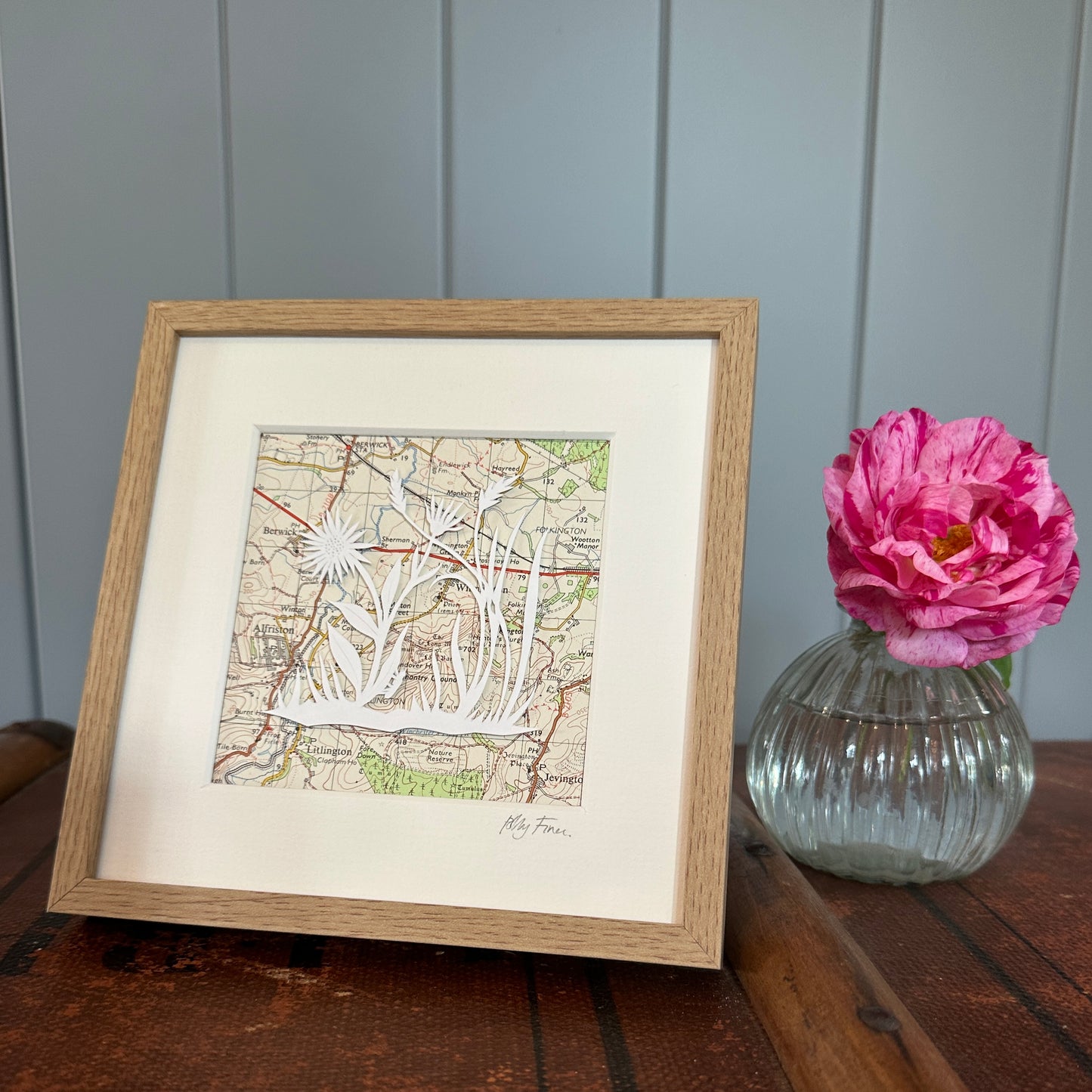 A group of flowers and grasses over an OS map extract in a wood effect frame next to a flower for scale