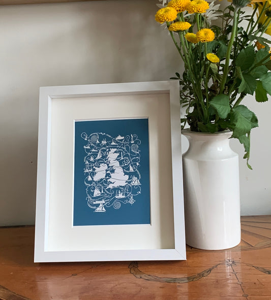An image of a papercut  map of UK and shipping forecast names, white on a dark turquoise background in a white frame on a tabletop.
