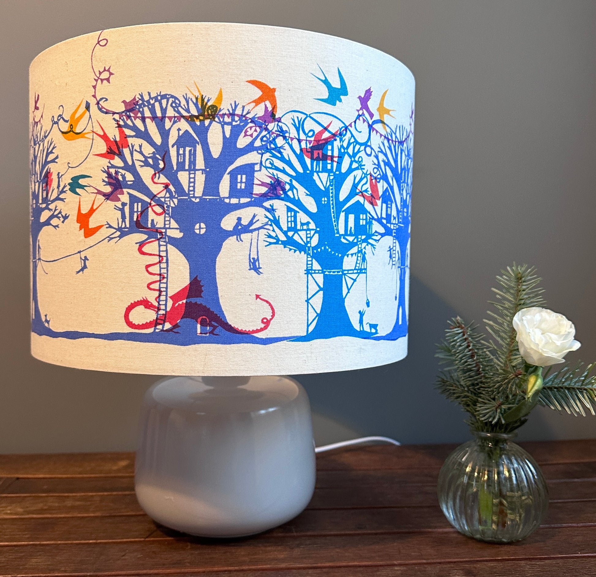 Table lampshade with blue and turquoise  trees with treehouses on cream background with multi coloured birds, bunting and a pink dragon.