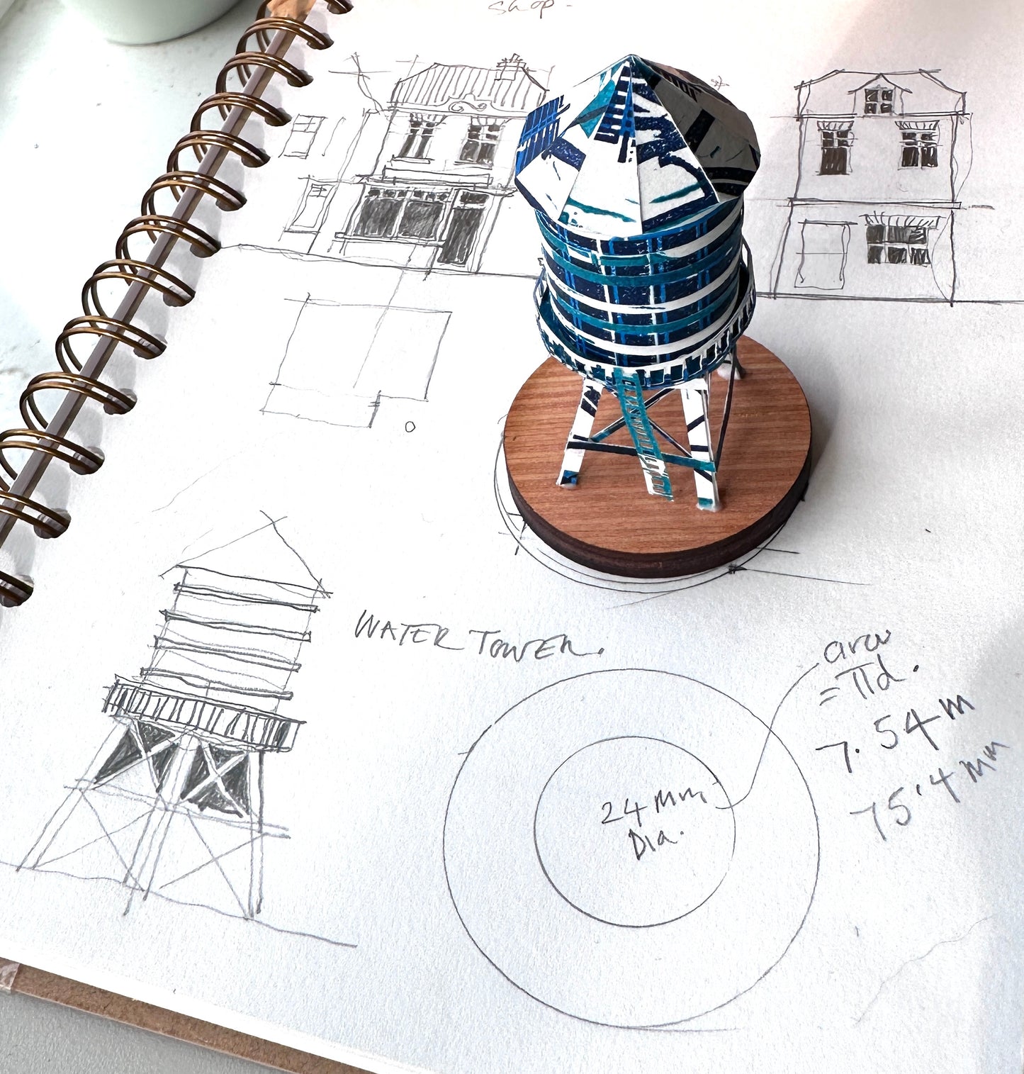 Handcut paper water tower over printed in shades of blue on wood base sitting on original sketch of water tower 