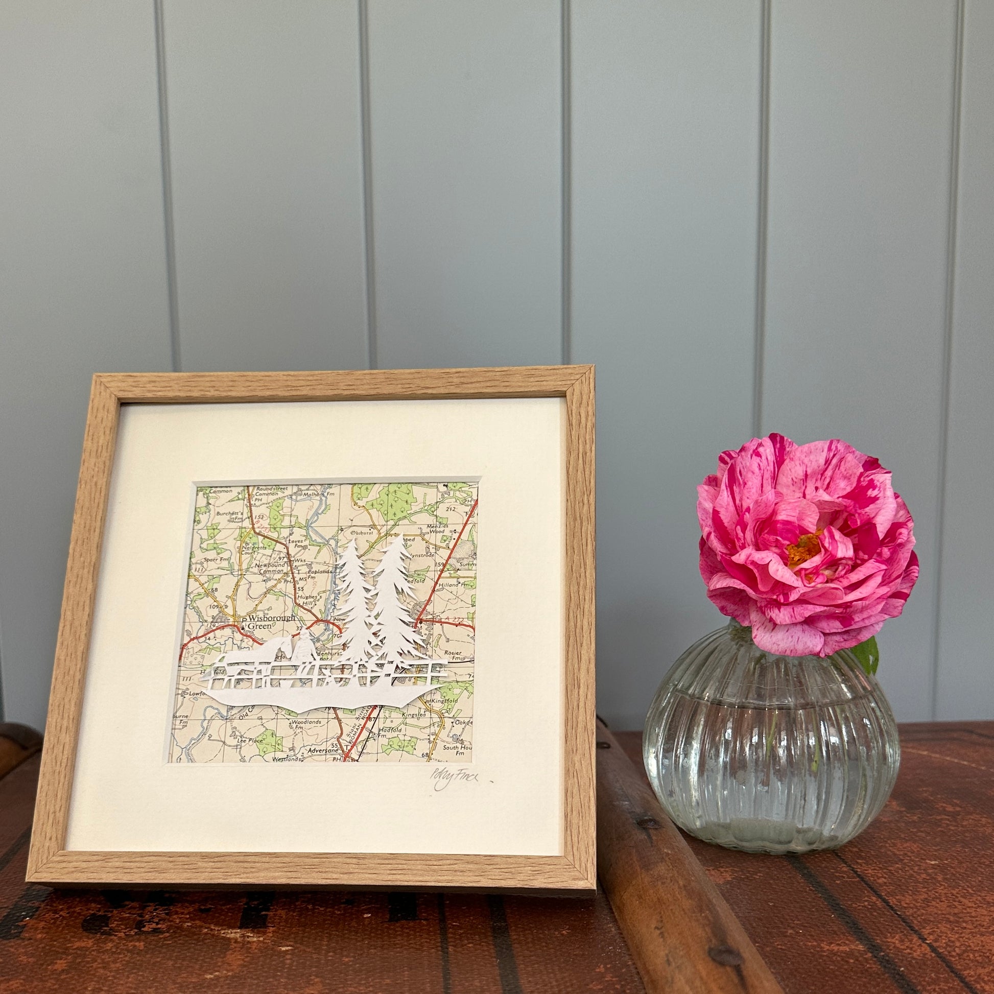 Papercut of a horse, a girl and dog next to two tall fir trees in wood effect frame next to flower for scale