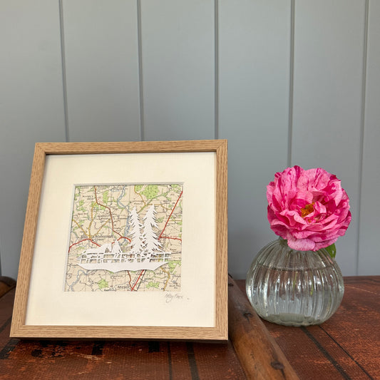 Papercut of a horse, a girl and dog next to two tall fir trees in wood effect frame next to flower for scale