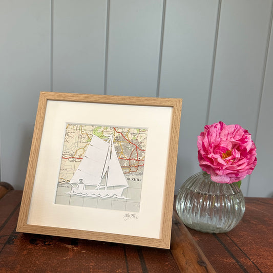 papercut of gaff rigged sailing boat on OS map in wood effect frame next to flower for scale