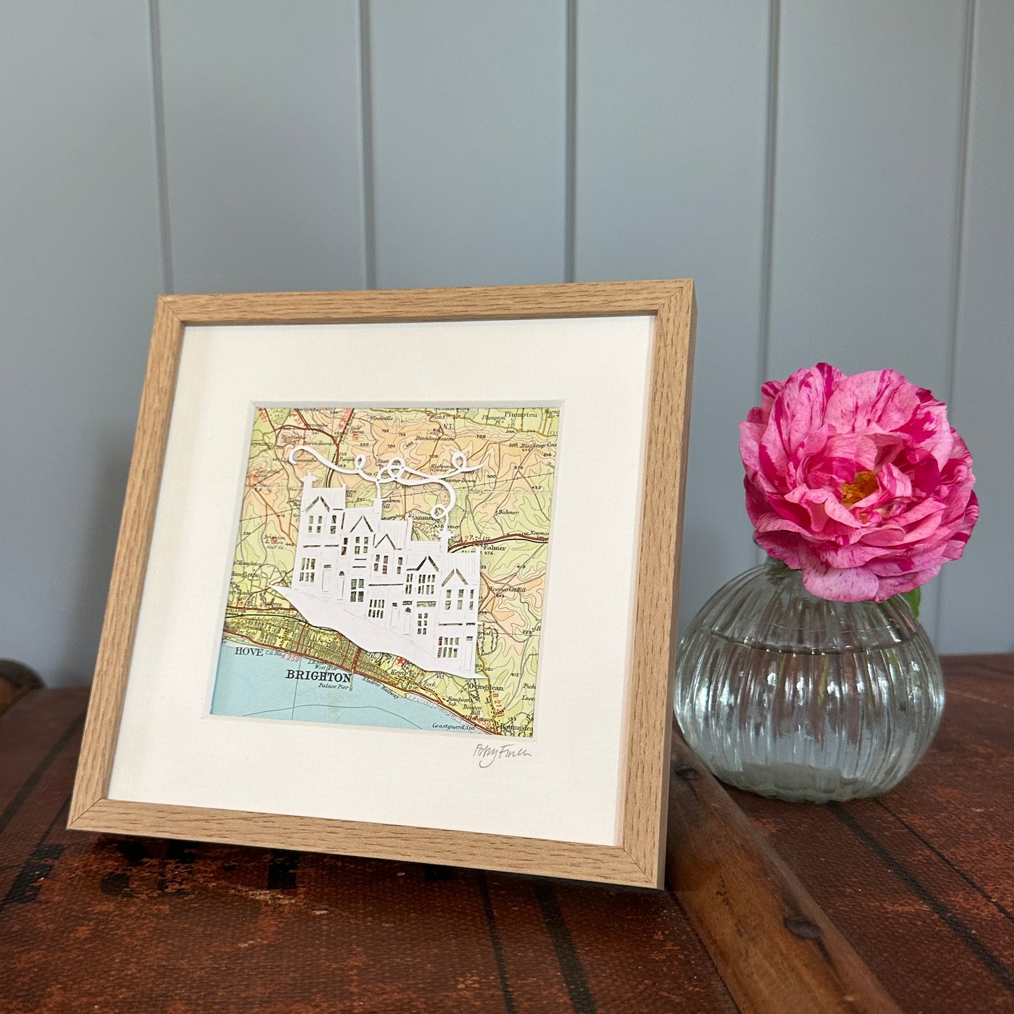 papercut of 5 houses on hillside on OS map extract in wood effect frame next to flower for scale
