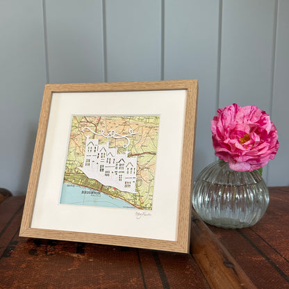 papercut of 5 houses on hillside on OS map extract in wood effect frame next to flower for scale
