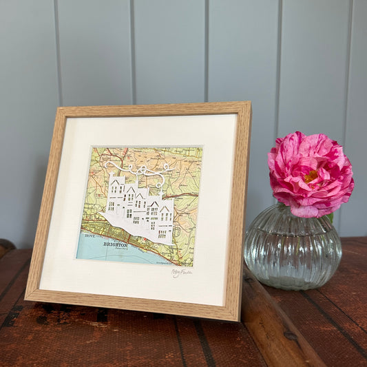 papercut of 5 houses on hillside on OS map extract in wood effect frame next to flower for scale