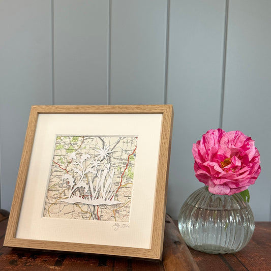 Papercut of a thistle and flowers over and OS map extract in a wood effect frame next to flower for scale